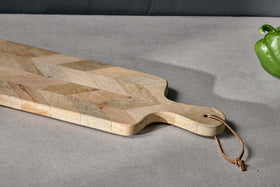 The nkuku Nalbari Chopping Board - Natural, crafted from sustainable mango wood with a chevron pattern and leather loop handle, rests on a light gray countertop. A green bell pepper is in the upper right; the background wall is dark gray.