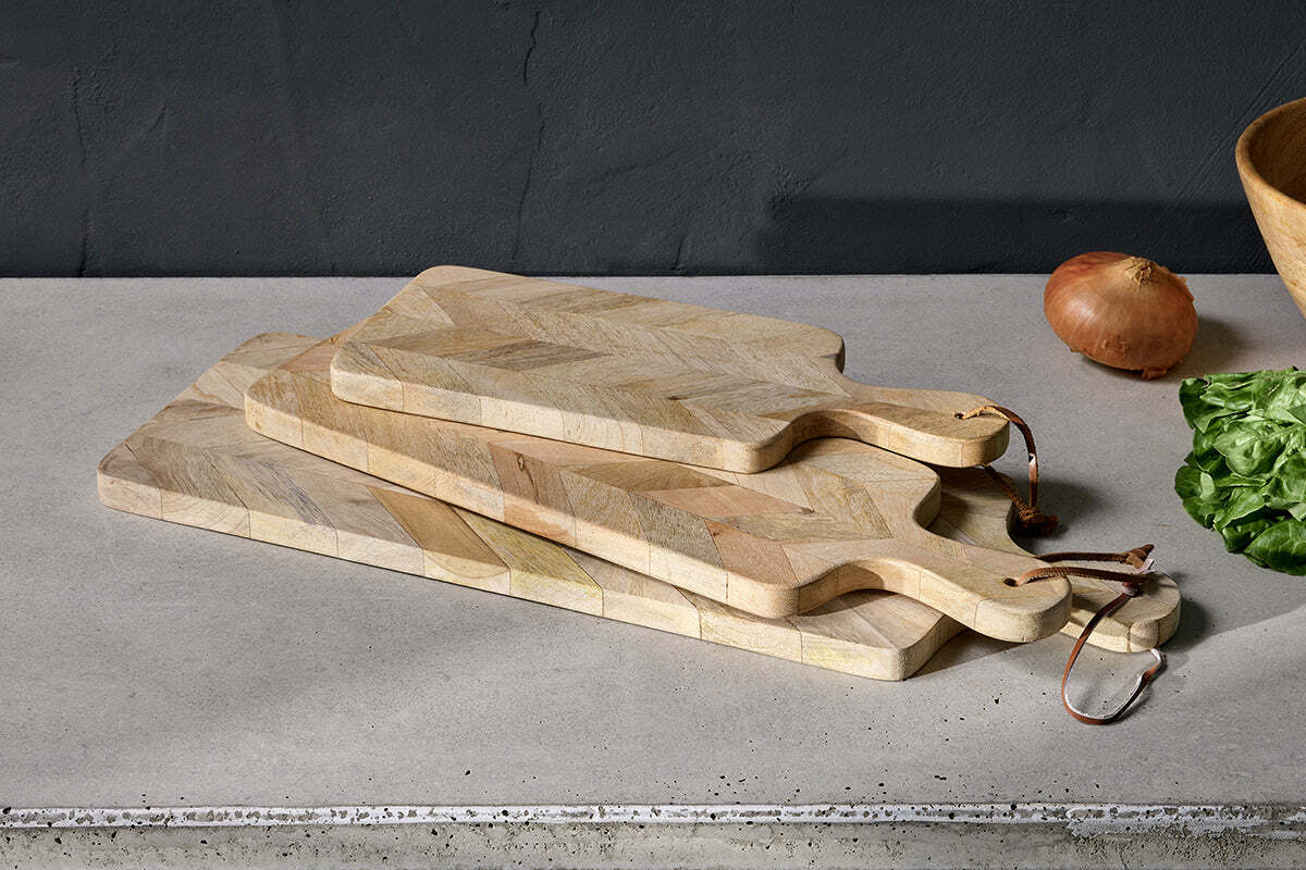 Three light Nkuku Nalbari Chopping Boards in natural mango wood are stacked on a gray countertop. Behind them are a brown onion, a wooden bowl, and leafy greens set against a dark, textured wall.