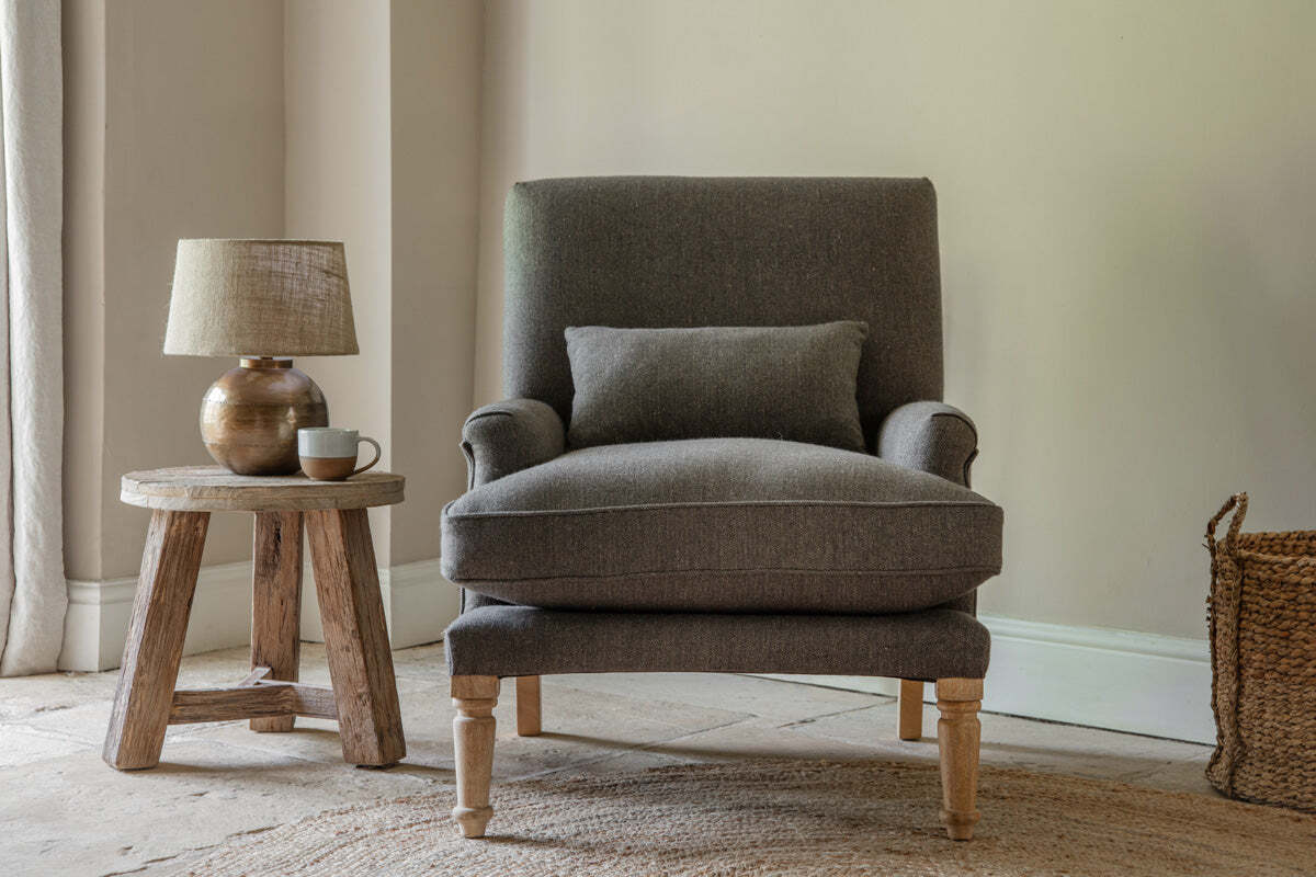 A cozy corner features the nkuku Nalla Wool Armchair in charcoal, accented by a herringbone cushion. The chair sits on a woven rug beside a rustic wooden side table with a ceramic lamp and cup, bathed in soft natural light through curtains.