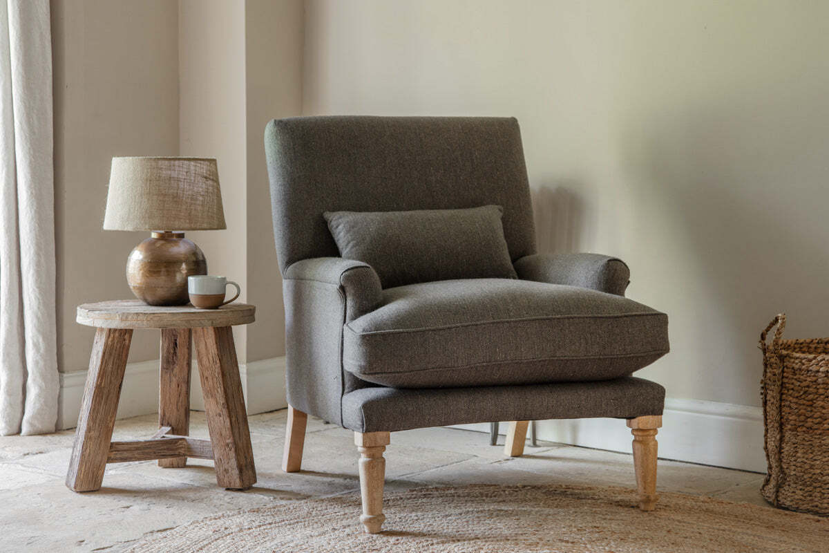 The nkuku Nalla Wool Armchair in Charcoal, complete with a cushion, sits on a woven rug by a beige wall. Next to it, a rustic wooden side table holds a fabric-shade lamp and white mug, while soft light reveals part of a woven basket.