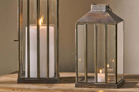 Two nkuku Nampo Lanterns - Aged Antique, made of brass and glass, sit on wood. The left contains a large white pillar candle with a tall flame; the right holds a smaller lit candle, both casting a warm glow against a neutral background.