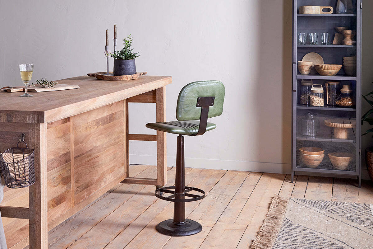 A modern kitchen features a wooden table, the nkuku Narwana Bar Chair in rich green leather with a metal base, a glass of white wine, potted plants, and a tall glass cabinet with dishes. Light wood floors and neutral walls complete the look.