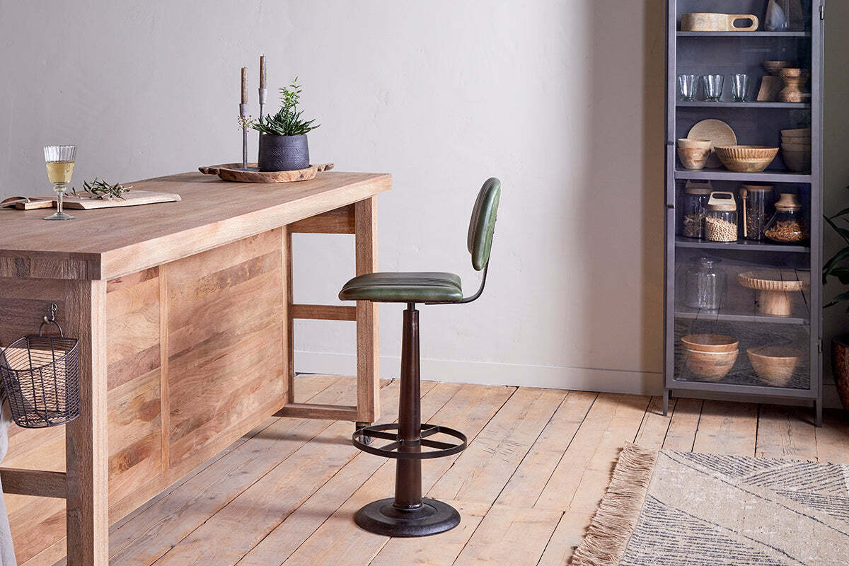The Narwana Bar Chair - Rich Green by nkuku features green leather and a dark metal pedestal base, paired with a rustic wood island. Open metal shelves hold bowls and jars; light wood floors and neutral walls enhance the industrial feel.