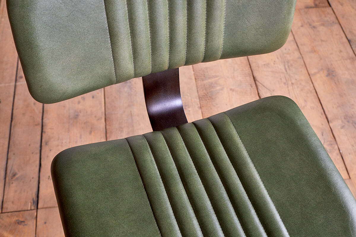 Two Narwana Bar Chairs in rich green by nkuku are shown from above. A dark metal support connects the industrial-style seats, which rest on a wooden floor for a modern bar stool effect.
