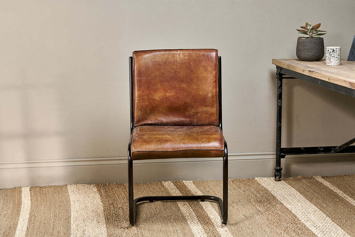 The Narwana Leather Desk Chair - Aged Tan by nkuku features a brown leather seat and backrest on a sleek iron frame. It stands on a beige woven rug beside a wooden desk with black metal legs and two small potted plants.