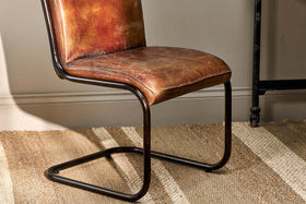 The Narwana Leather Desk Chair - Aged Tan by nkuku, with its worn brown leather seat and black iron frame, sits atop a woven rug. Sunlight casts shadows on the chair, highlighting its vintage style against a beige wall.