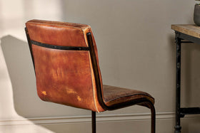 The Narwana Leather Desk Chair - Aged Tan by nkuku sits beside a wooden table with a black metal frame, sunlight highlighting the chair’s aged leather texture and iron backrest.