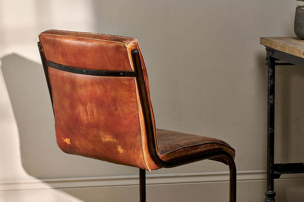 The Narwana Leather Desk Chair - Aged Tan by nkuku sits beside a wooden table with a black metal frame, sunlight highlighting the chair’s aged leather texture and iron backrest.