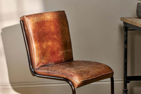 A close-up of the Narwana Leather Desk Chair - Aged Tan by nkuku showcases its vintage-style distressed brown leather, curved iron frame, and padded seat and backrest—an ideal piece to elevate any interior space.