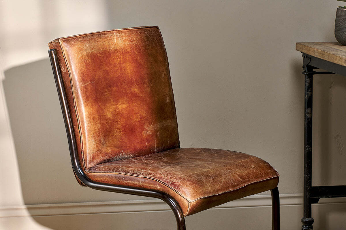 A close-up of the Narwana Leather Desk Chair - Aged Tan by nkuku showcases its vintage-style distressed brown leather, curved iron frame, and padded seat and backrest—an ideal piece to elevate any interior space.