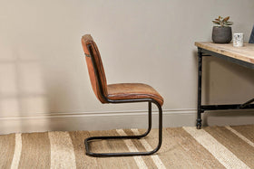 The Narwana Leather Desk Chair in aged tan by nkuku, with an iron frame, sits on a beige woven rug beside a wooden desk with black metal legs. The desk holds a small potted succulent and white patterned cup, set against a light gray wall for a calm look.