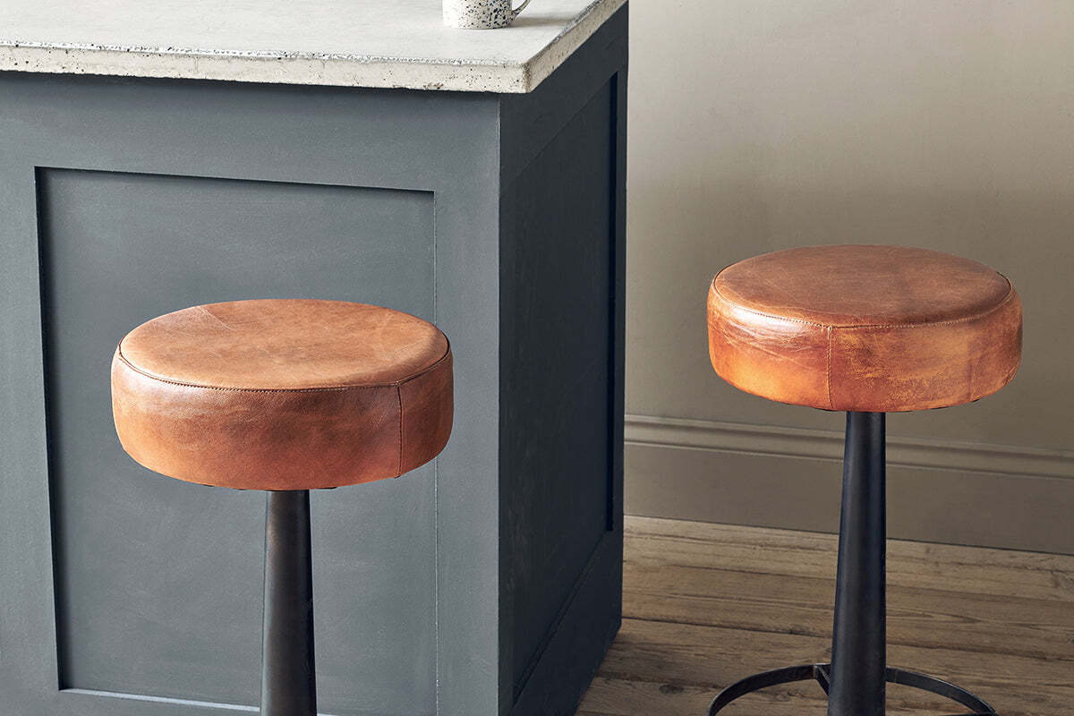 Two Nkuku Narwana Leather Round Stools in aged tan with black metal bases are set by a gray paneled kitchen island with a white countertop, on light wood flooring beside a beige wall.