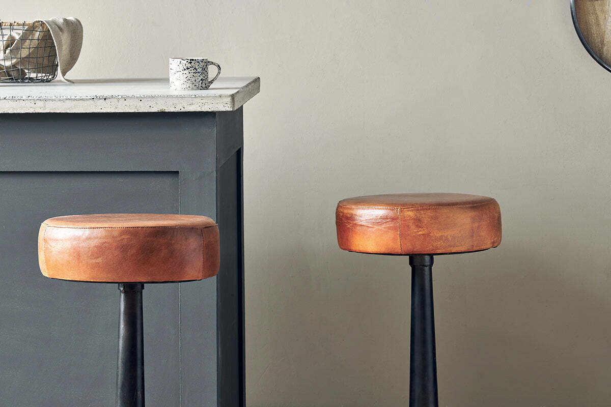 Two nkuku Narwana Leather Round Stools in aged tan, each with round seats and black metal legs, stand by a gray kitchen counter topped with a speckled mug and small basket. The background wall is light beige.