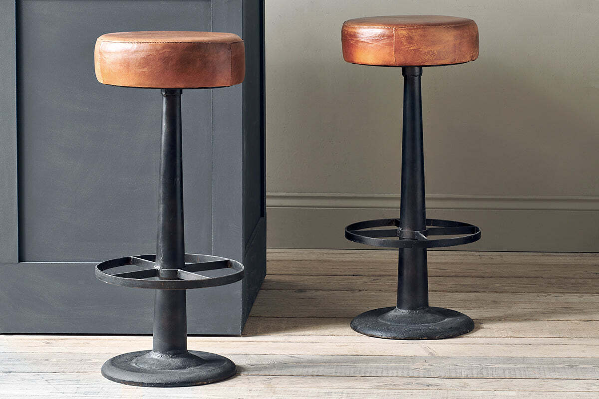 Two nkuku Narwana Leather Round Stools in Aged Tan, featuring round leather seats, black metal bases with footrests, and a sturdy minimalist design, stand on a light wooden floor by a gray paneled counter.