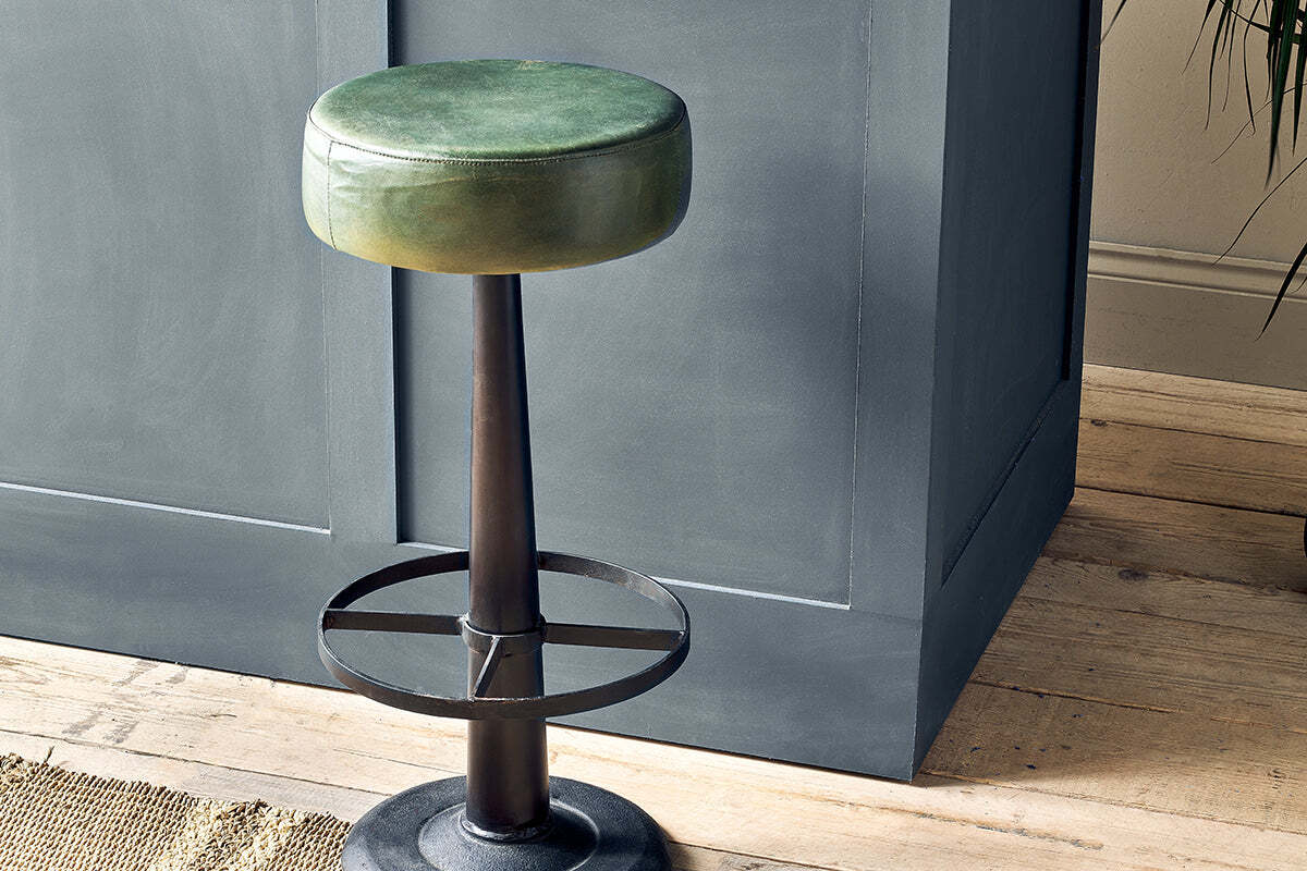 The nkuku Narwana Leather Round Stool in Rich Green, featuring a black metal base and circular footrest, stands on light wooden flooring next to a blue-gray paneled kitchen island. A woven rug and plant are partially visible nearby.