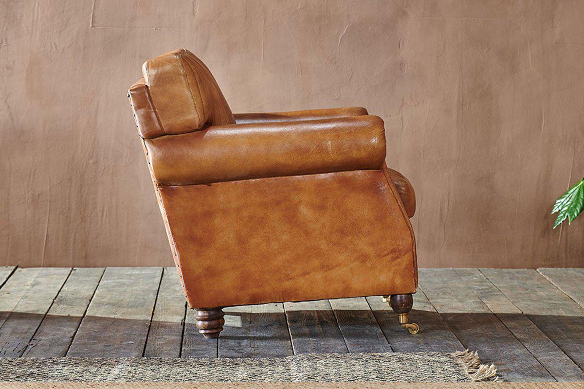 The nkuku Narwana Ribbed Leather Armchair in Aged Tan is showcased in profile on a rustic wooden floor, set against a textured brown wall with a leafy green plant partially visible to the right.