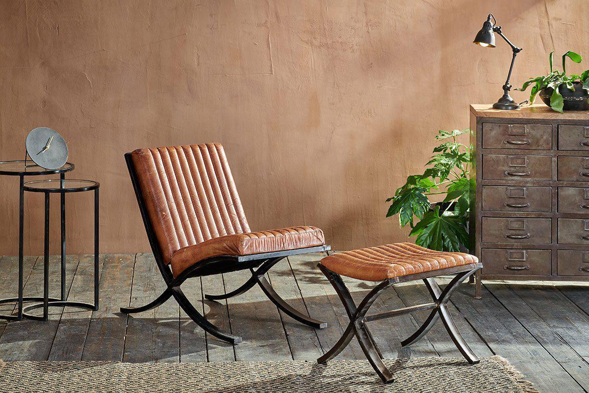 A modern living space features a brown leather chair and ottoman on dark wood floors. The nkuku Narwana Ribbed Leather Footstool in Aged Tan adds comfort and style, complemented by leafy plants, a chest of drawers, and a textured rug.
