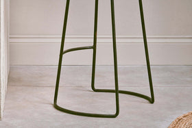 Close-up of the dark metal base and thin, curved legs with a support bar on the nkuku Narwana Ribbed Leather Stool - Aged Tan - Large, set against a textured, light-colored wall with molding in the background.