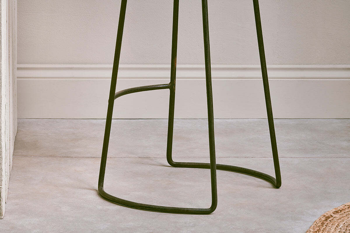 Close-up of the dark metal base and thin, curved legs with a support bar on the nkuku Narwana Ribbed Leather Stool - Aged Tan - Large, set against a textured, light-colored wall with molding in the background.
