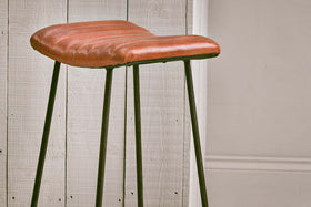 The Narwana Ribbed Leather Stool - Aged Tan (Large) by nkuku features a ribbed leather seat and slim black metal legs, adding a stylish mix of textures and tones to your minimal space against white paneling and beige wainscoting.