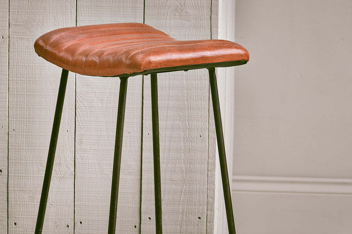 The Narwana Ribbed Leather Stool - Aged Tan (Large) by nkuku features a ribbed leather seat and slim black metal legs, adding a stylish mix of textures and tones to your minimal space against white paneling and beige wainscoting.
