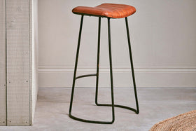The Narwana Ribbed Leather Stool in Aged Tan (Large) by nkuku, featuring a padded seat and dark metal base, sits in front of a pale gray wall.