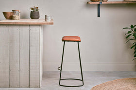 A minimalist kitchen with a wooden countertop, small potted plants, and jars on shelves. The nkuku Narwana Ribbed Leather Stool in Aged Tan (Large) with black metal legs sits on the gray floor. A jute rug and leafy plant are partially visible in front.