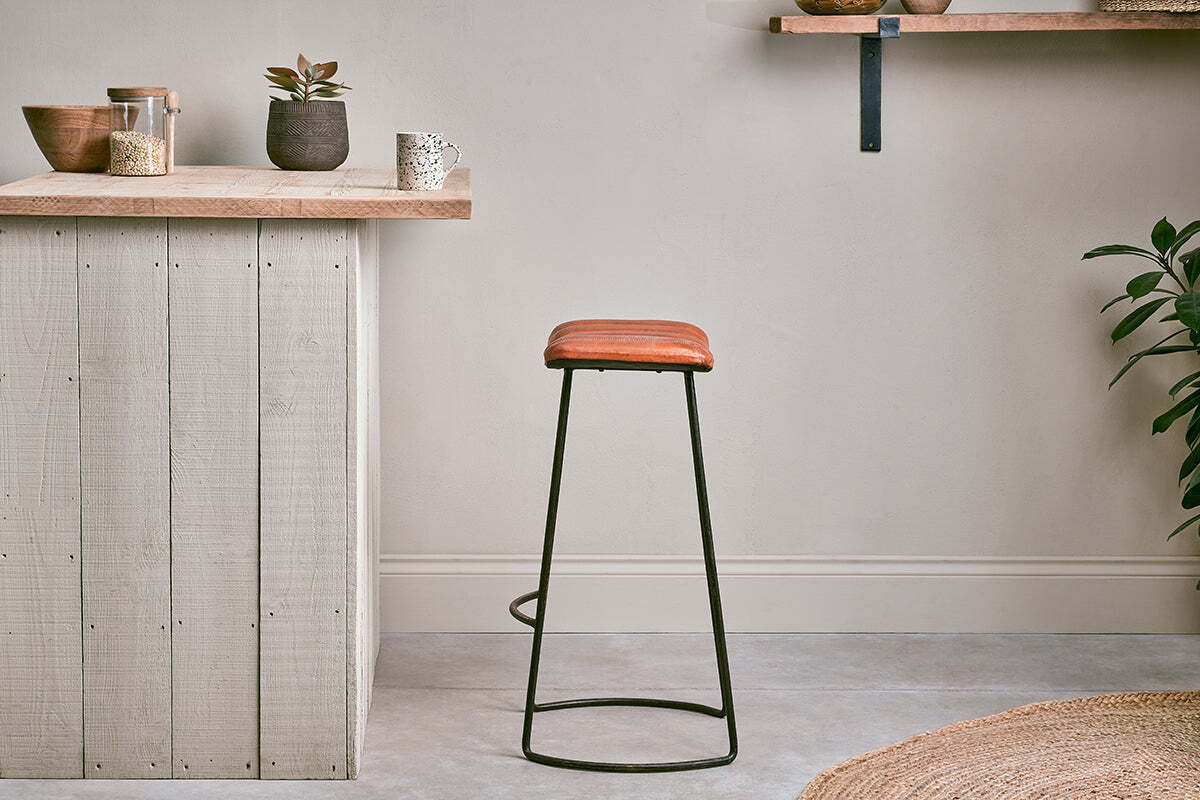 A minimalist kitchen with a wooden countertop, small potted plants, and jars on shelves. The nkuku Narwana Ribbed Leather Stool in Aged Tan (Large) with black metal legs sits on the gray floor. A jute rug and leafy plant are partially visible in front.