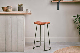 A minimalist kitchen features the nkuku Narwana Ribbed Leather Stool in Aged Tan (Large) with thin black metal legs. A wood countertop, white panelled bar, and shelves with plants create a cozy, modern feel.