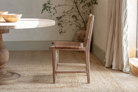 The nkuku Nayan Leather & Jute Dining Chair in tan sits beside a round mango wood table on a woven rug, sunlight pouring through light curtains, with a potted plant featuring long branches in the background.