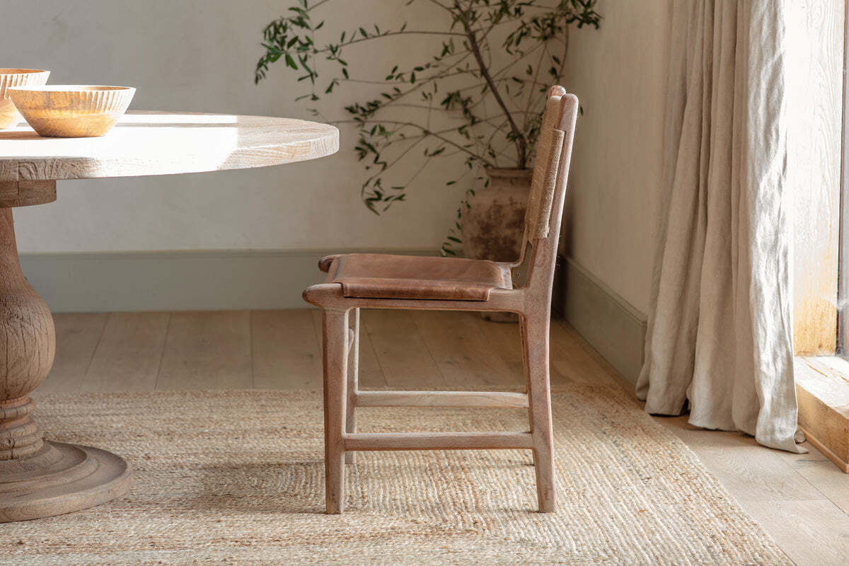 The nkuku Nayan Leather & Jute Dining Chair in tan sits beside a round mango wood table on a woven rug, sunlight pouring through light curtains, with a potted plant featuring long branches in the background.