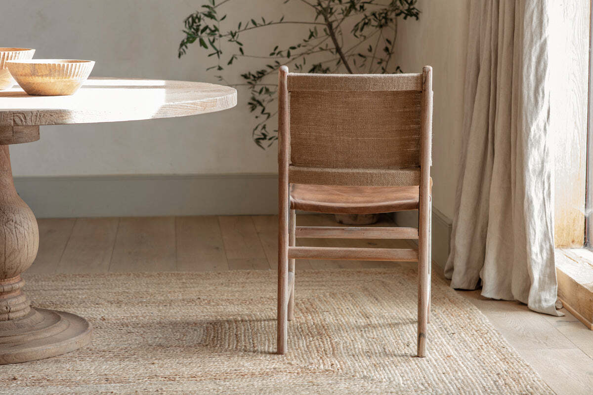 The Nayan Leather & Jute Dining Chair by nkuku, with its woven seat and back in tan, faces a round wooden table. Sunlight filters through sheer curtains onto a textured rug and pale wooden floor, with a leafy plant and bowls in the background.