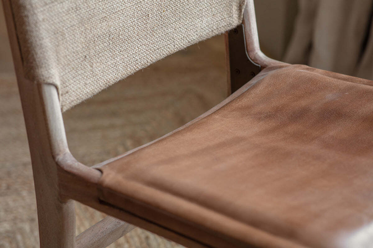 Close-up of the nkuku Nayan Leather & Jute Dining Chair - Tan, featuring a smooth buffalo leather seat, woven jute backrest, and sturdy mango wood frame against softly blurred flooring and light curtains in the background.
