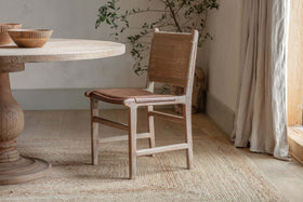 The Nayan Leather & Jute Dining Chair in tan by nkuku sits beside a round wooden table. Light walls, a woven rug, a corner plant, and sheer curtains complete the space’s cozy, natural vibe.