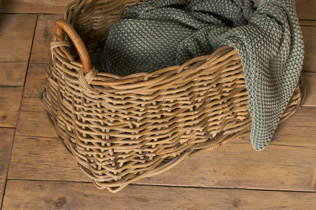 The Netra Rattan Basket - Natural by nkuku sits on a wooden floor, its sturdy handle and chunky-knit greenish-grey blanket draped over the edge bringing a cozy, homey feel to any space.