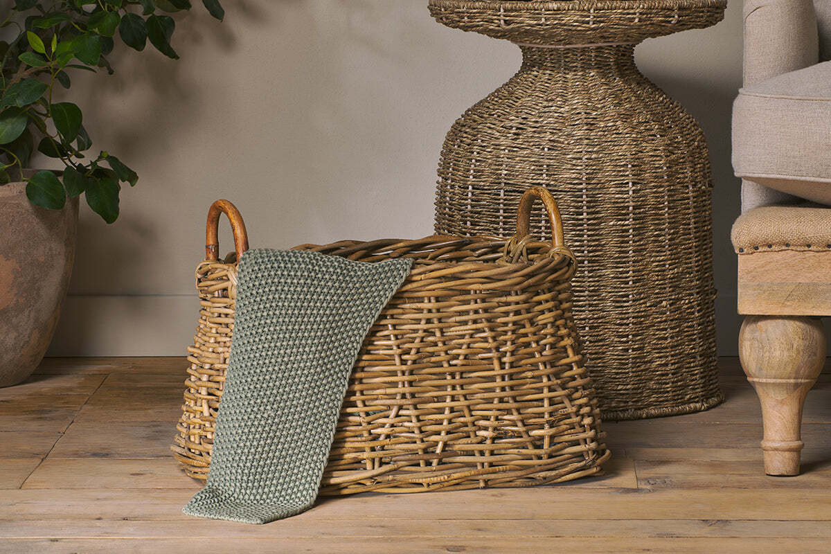 The Netra Rattan Basket - Natural by nkuku, featuring wooden handles, sits on a wooden floor with a green knit blanket. Behind the basket are a woven table, part of a beige upholstered chair, and a large potted plant.