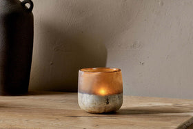 A small candle glows inside the Ngolo Tealight Holder - Antiqued Amber by nkuku, its hand-blown, partly frosted glass resting on a wooden surface against a beige wall. To the left, a dark, blurred vase appears in the background.