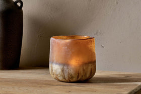 The Ngolo Tealight Holder - Antiqued Amber by nkuku rests on a wooden surface against a beige wall, its mottled amber glass casting a warm glow. On the left edge, a dark blurred vase is partly visible in the background.