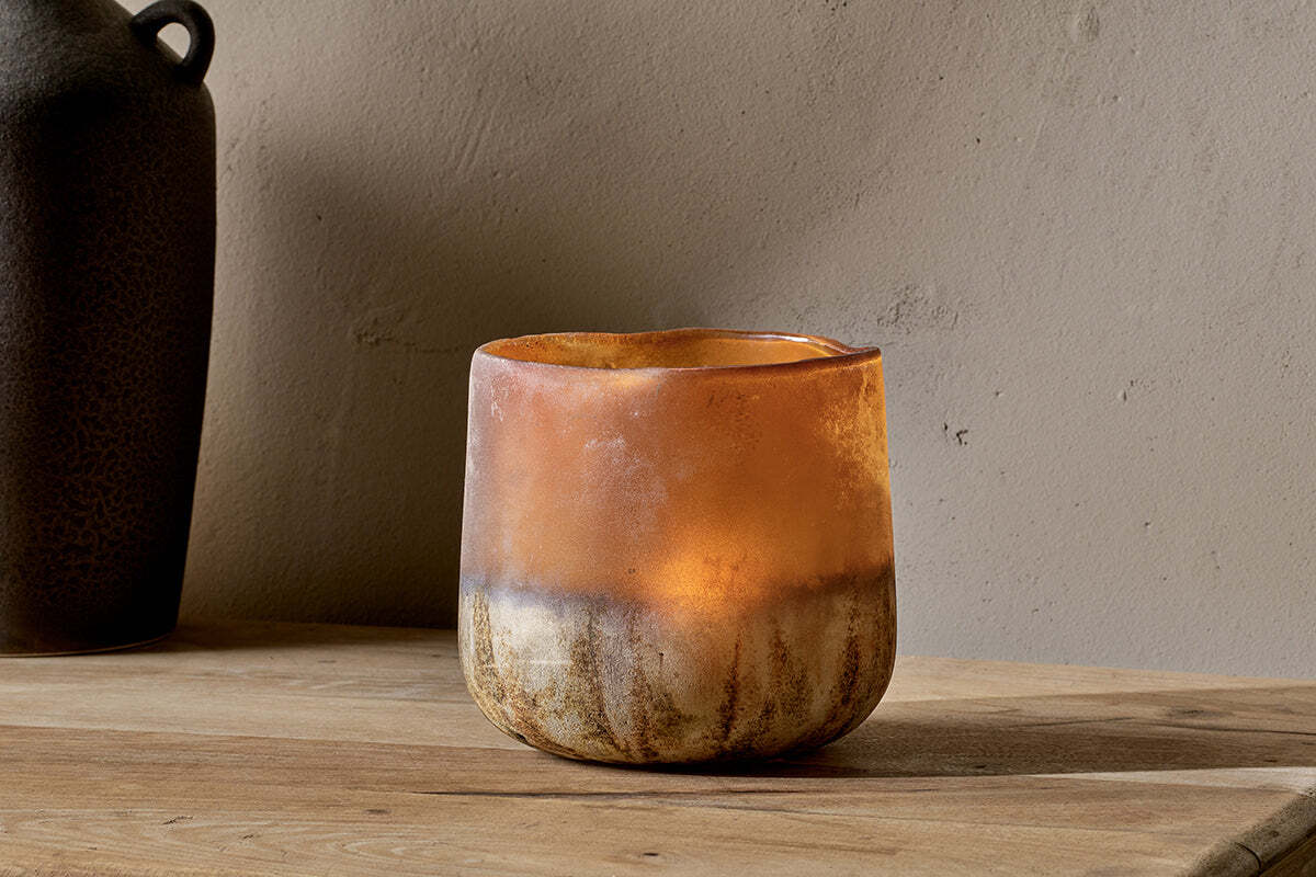 The Ngolo Tealight Holder - Antiqued Amber by nkuku rests on a wooden surface against a beige wall, its mottled amber glass casting a warm glow. On the left edge, a dark blurred vase is partly visible in the background.