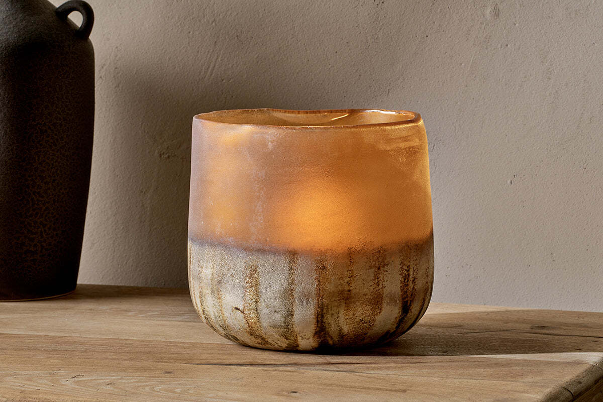 The nkuku Ngolo Tealight Holder - Antiqued Amber features a rustic, hand-blown round design with a frosted amber-to-brown gradient, softly glowing on a wooden surface against a textured beige wall.