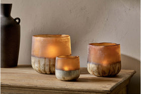 Three Ngolo Tealight Holders by nkuku in antiqued amber glow warmly on a wooden surface against a beige wall, while a dark matte vase in the background adds stylish contrast to this cozy, rustic scene.