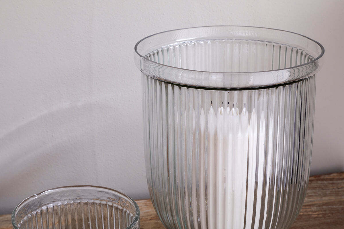 A close-up of nkuku's Nia Recycled Glass Candle Holders in clear, featuring a fluted design on wood—a larger holder with a white candle and the upper edge of the smaller empty holder visible against a plain light wall.