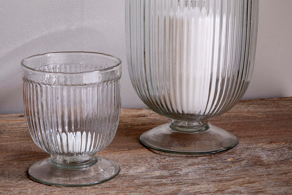 Two Nia Recycled Glass Candle Holders by nkuku, featuring a fluted design and flared bases, sit on a rustic wooden surface. The taller clear holder contains white powder; the smaller is empty. Both can also be used as unique goblets.