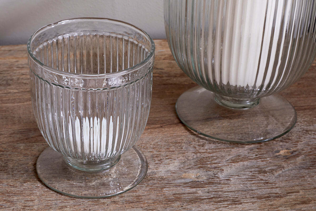 Two nkuku Nia Recycled Glass Candle Holders in clear, featuring a fluted design and wide bases, rest on rustic wood. One holds a small white candle and the other, partially visible, contains a tall white candle. A plain light wall is behind them.