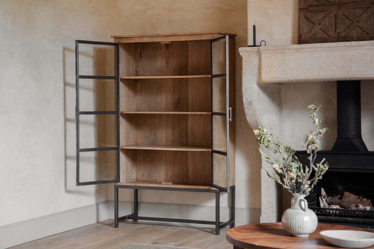 The Nirvi Wood & Iron Cabinet - Black by nkuku stands open beside a stone fireplace, showcasing its industrial style with black metal framing, glass doors, and three shelves. The cabinet rests on a light wooden floor near a round table with a leafy vase.