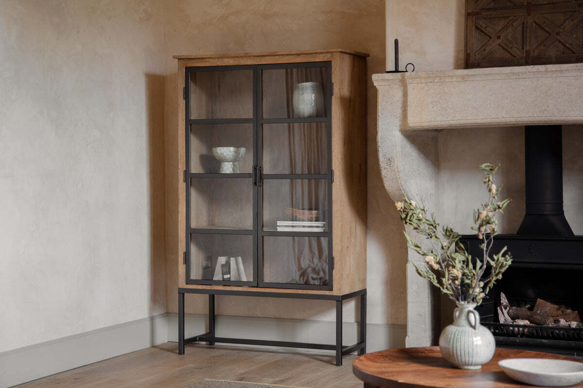 The nkuku Nirvi Wood & Iron Cabinet - Black stands in a neutral room by a stone fireplace, its wooden and glass panels showcasing bowls and books. A round table with a vase of leafy branches sits on the light wood floor in front.