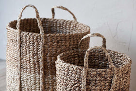 Two nkuku Nkomi Baskets, hand woven from natural textured fibers, sit side by side on a light floor. Both cylindrical storage baskets feature sturdy handles; the larger one is behind the smaller, set against a pale wall.