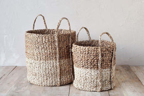 The Nkomi Baskets by nkuku are handwoven from natural fibers in beige and light brown. These cylindrical baskets, available in two sizes with handles, offer versatile storage and style for your home.