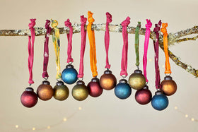 A mossy branch displays 12 nkuku Oko Mini Baubles - Mixed Matt in gold, blue, green, and red on fabric ribbons, with soft string lights glowing softly behind.