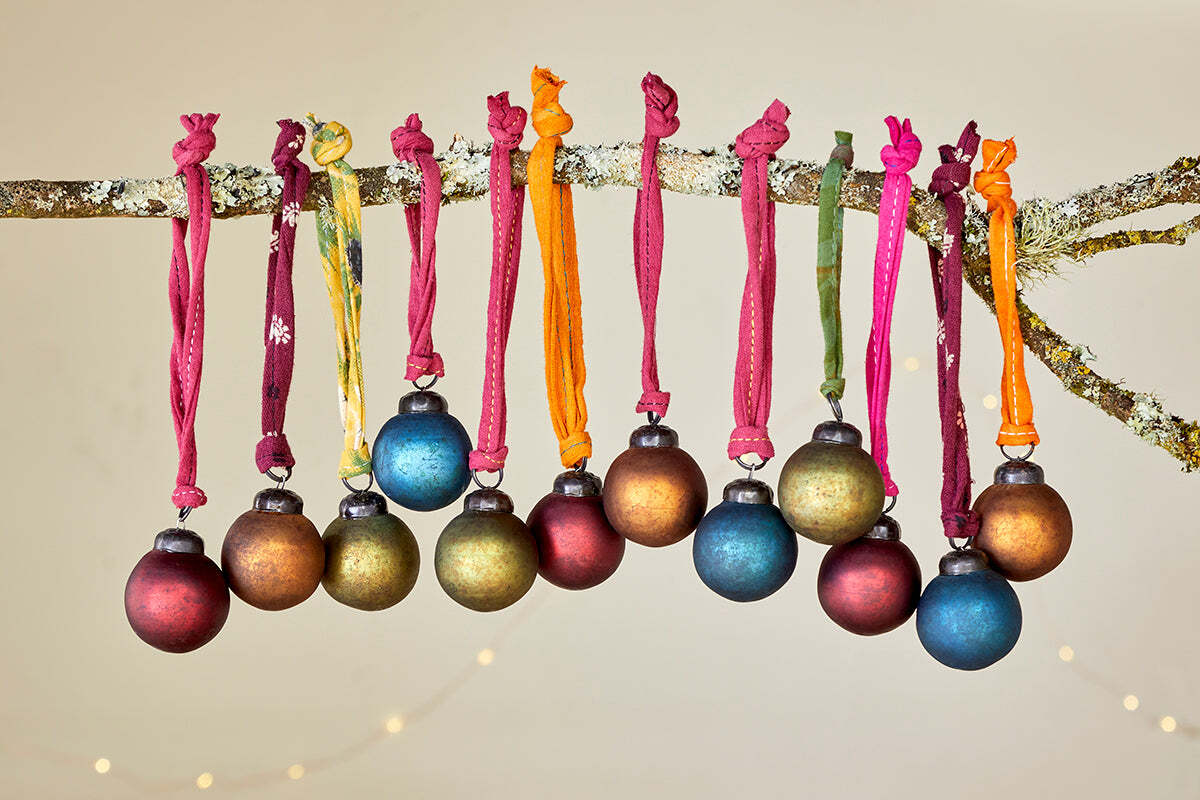 A mossy branch displays 12 nkuku Oko Mini Baubles - Mixed Matt in gold, blue, green, and red on fabric ribbons, with soft string lights glowing softly behind.
